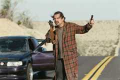 Leonardo DiCaprio stands in a road in his bathrobe holding up a rifle in One Battle After Another